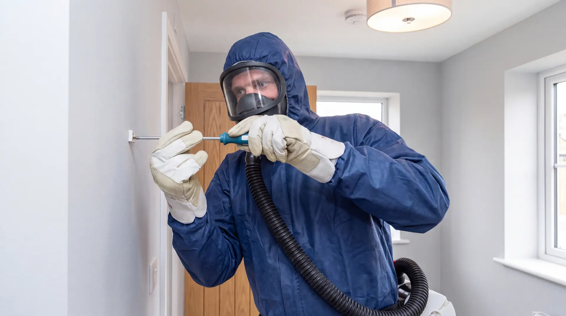 Fachperson im blauen Schutzanzug mit Vollmaske entnimmt eine staubarme Asbestprobe an einer Wand