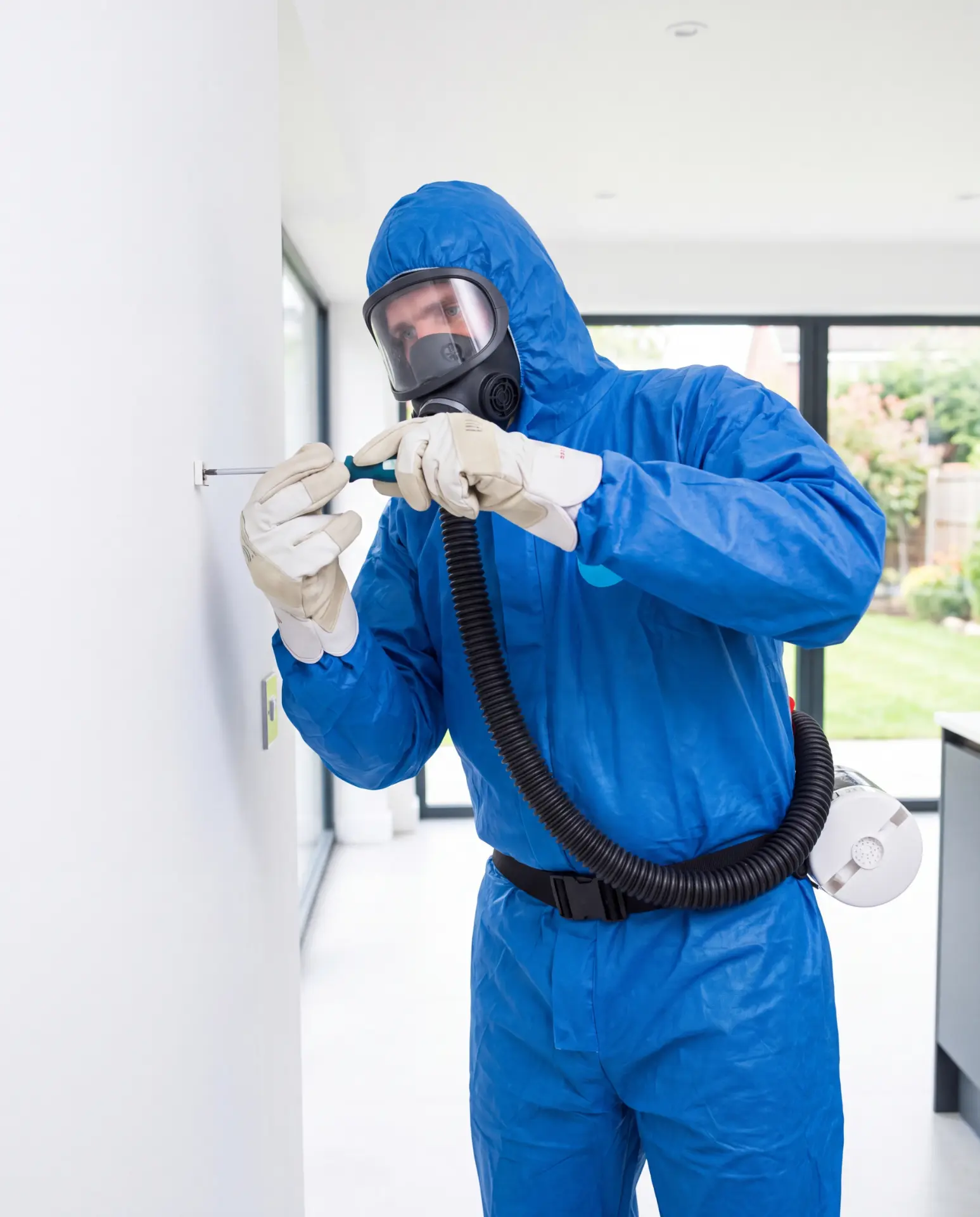 Fachperson im blauen Schutzanzug mit Vollmaske entnimmt staubarm eine Asbestprobe an einer Wand