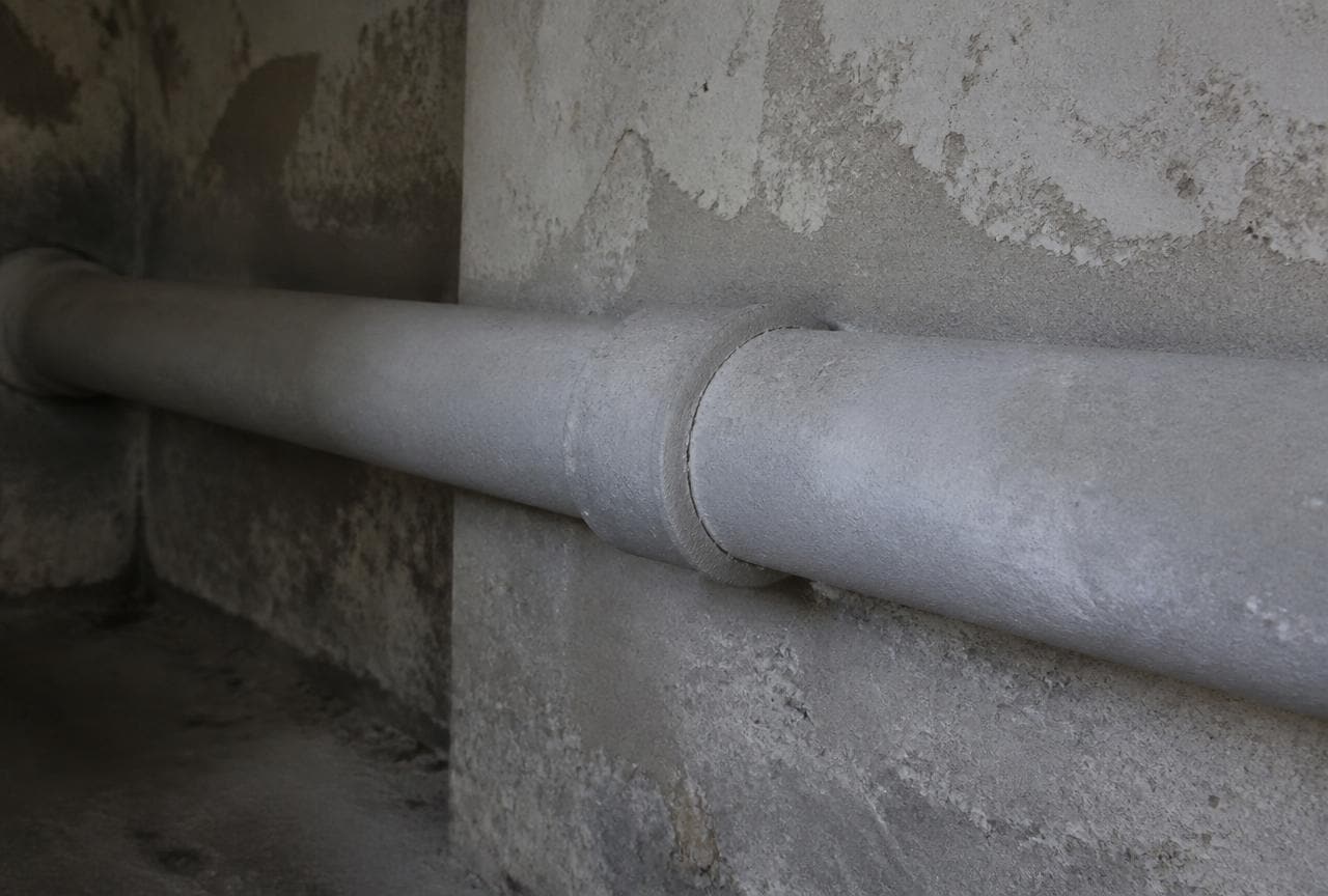 Graues Asbestzementrohr mit aufgeschobener Muffe an einer Kellerwand eines Altbaus