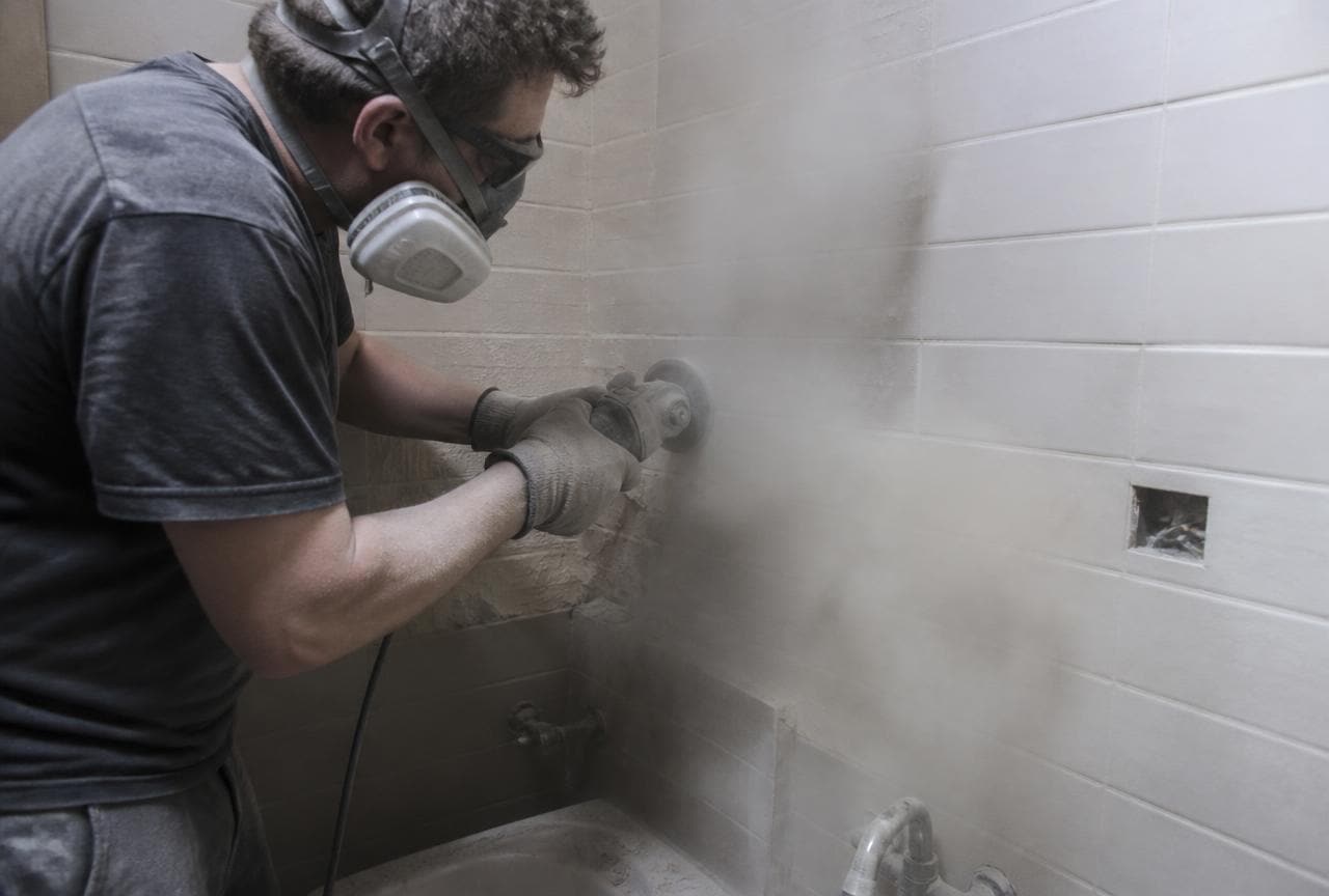 Handwerker flext eine Fliesenwand, dichte Staubwolke im kleinen Badezimmer