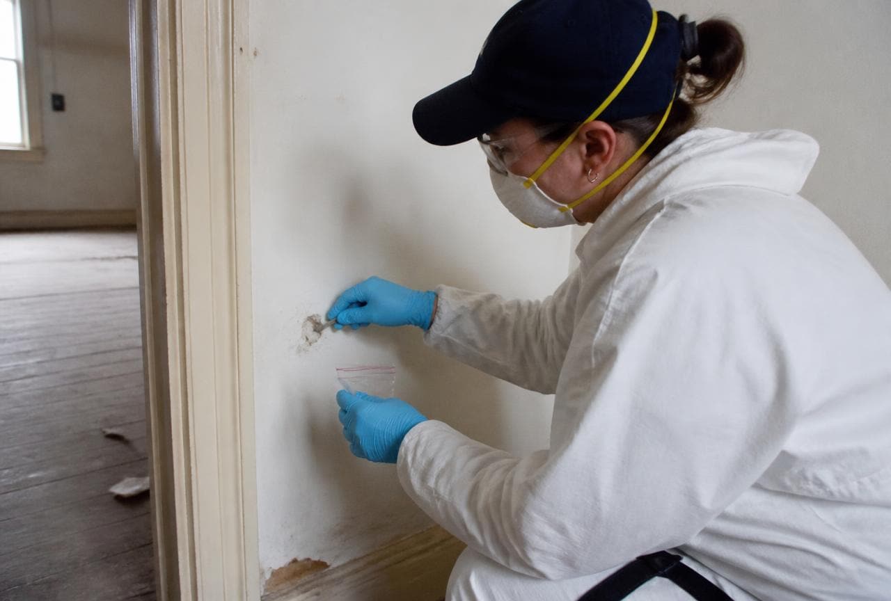 Fachperson in Schutzkleidung entnimmt eine Materialprobe aus einer Wand im leeren Haus