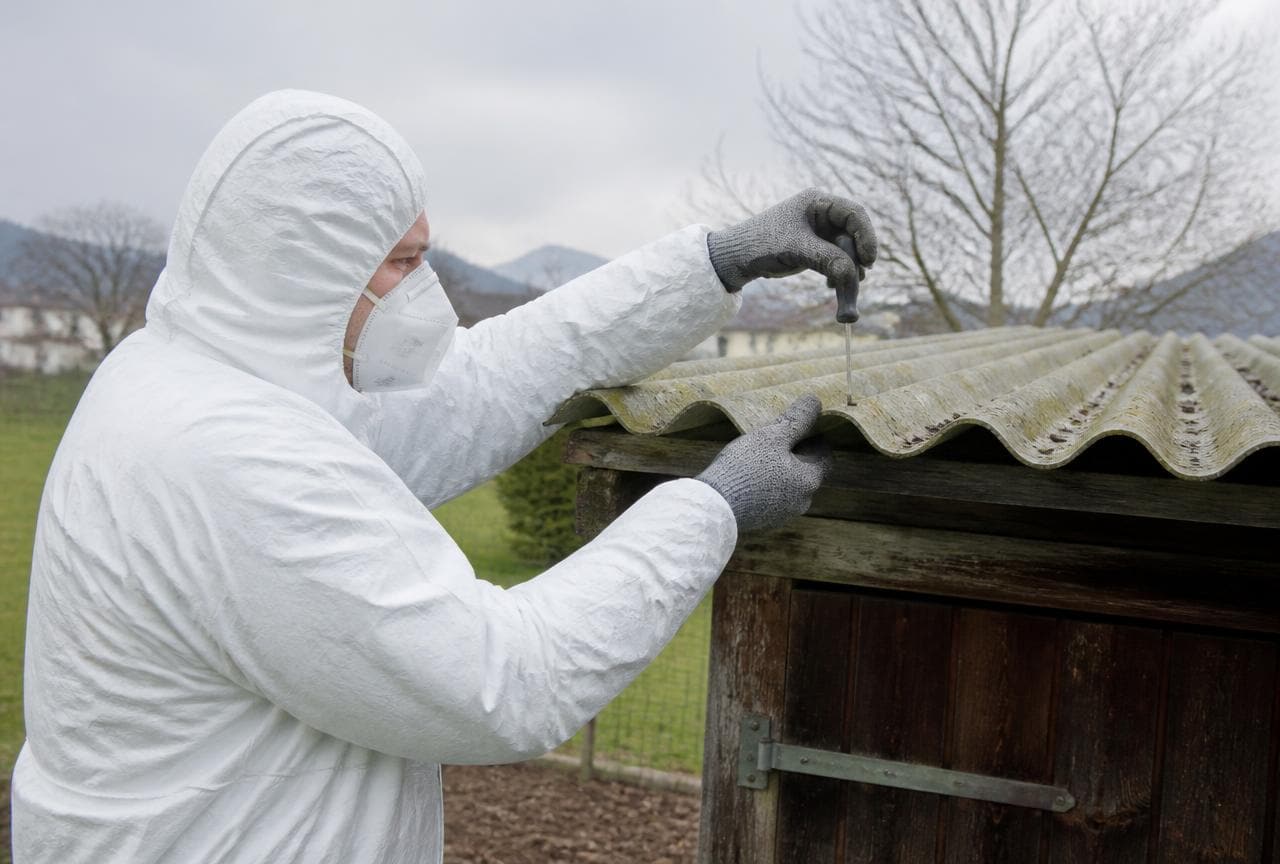 Person im Schutzanzug mit FFP3-Maske schraubt eine intakte Asbestzement-Wellplatte von einem Gartenhaus ab