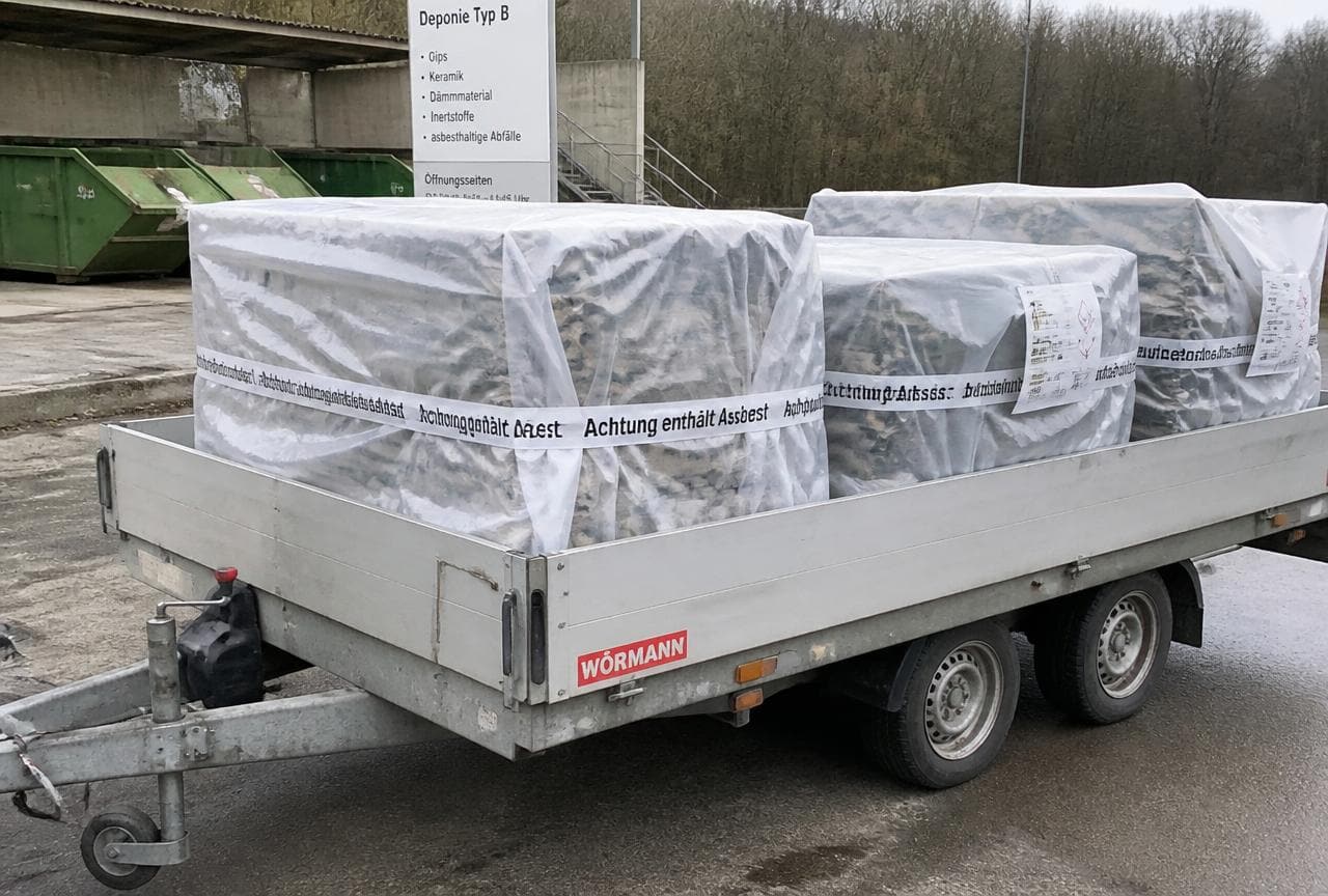 Doppelt verpackte und beschriftete Asbestplatten auf einem Anhänger vor einer Deponie