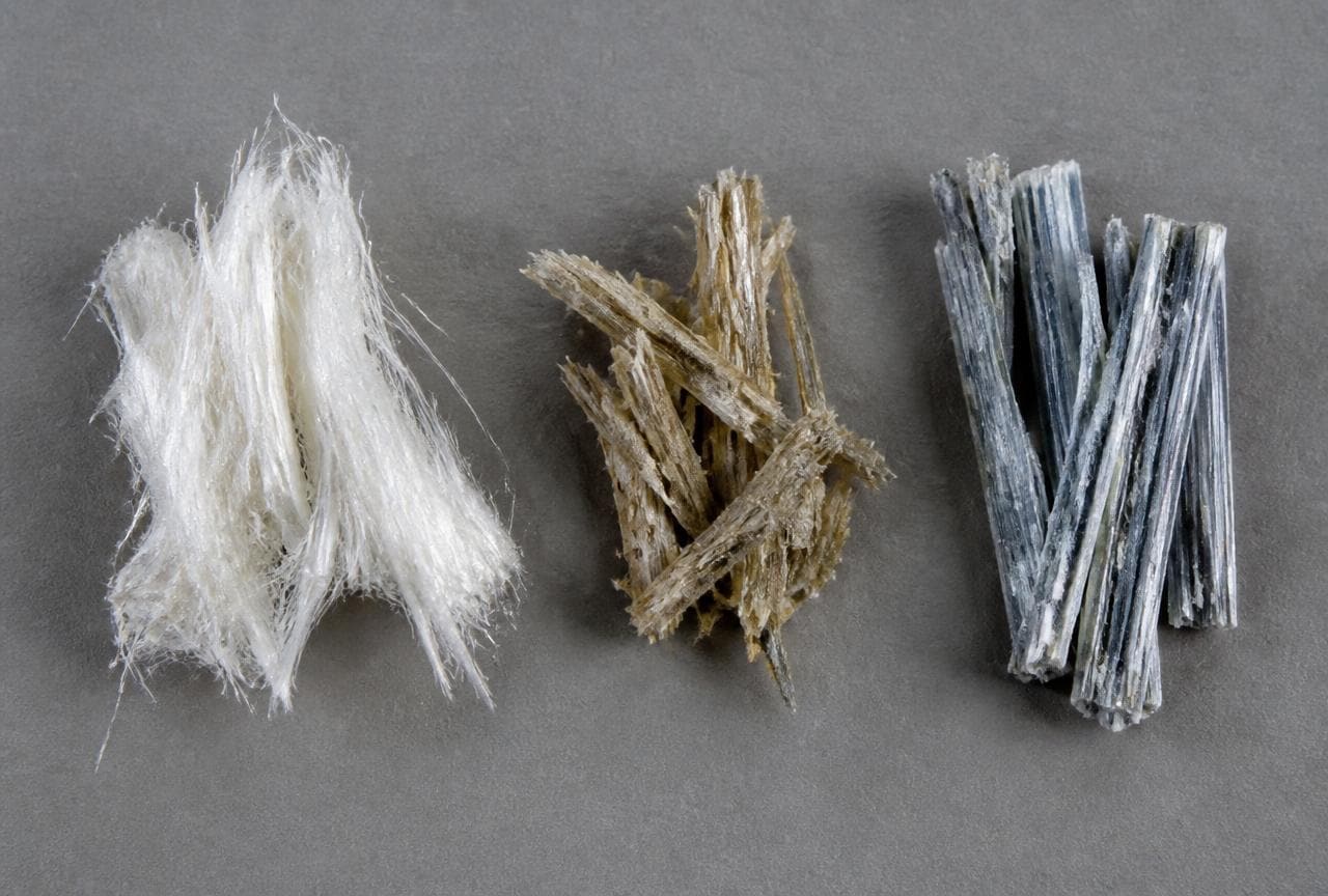 Weisse, braune und blaue Asbestfasern nebeneinander als Mineralproben auf grauem Untergrund